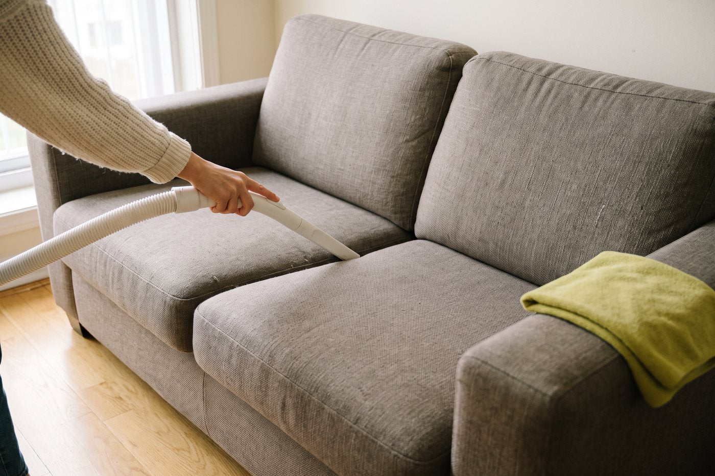 Eine Hand saugt mit der Fugendüse die Ritze zwischen zwei Sitzkissen eines grauen Stoffsofas ab