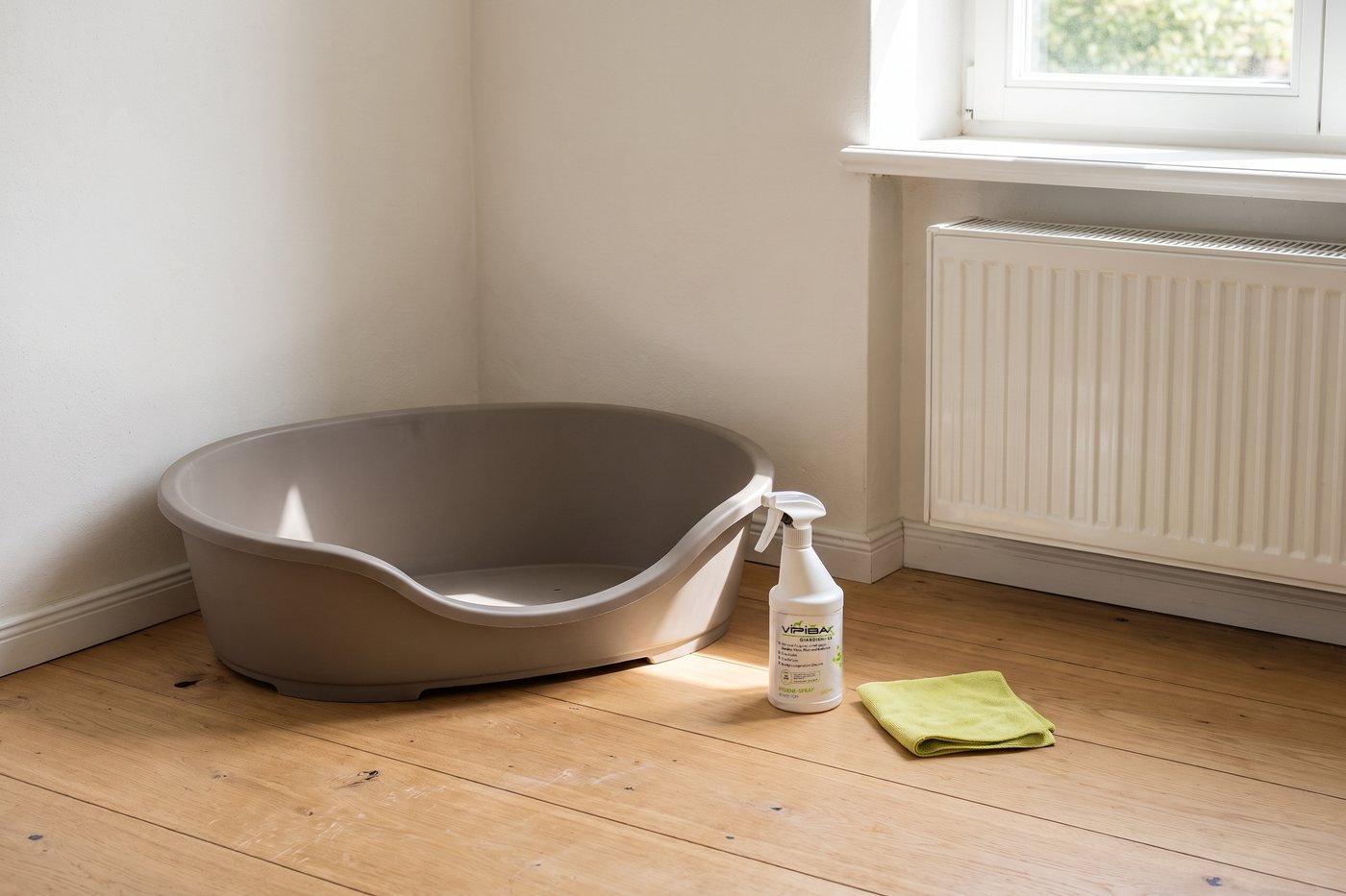 Leeres Kunststoff-Hundekörbchen in einer Zimmerecke, daneben das ViPiBaX Hygiene-Spray 500 ml und ein gefaltetes Tuch